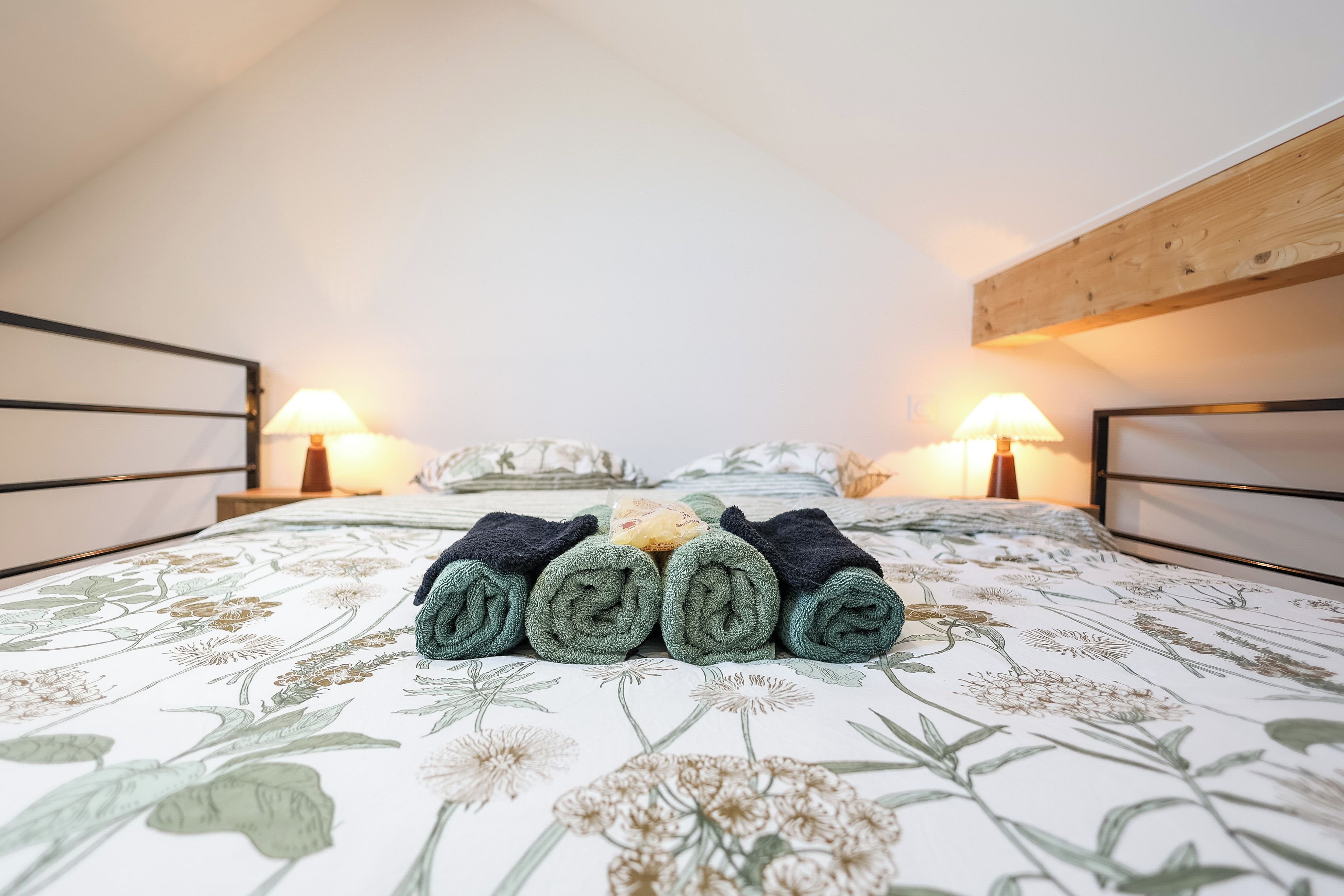 Een slaapkamer met een bloemenbedekking, een net opgemaakt bed met opgerolde handdoeken, twee bedlampen en een houten hoofdbord.