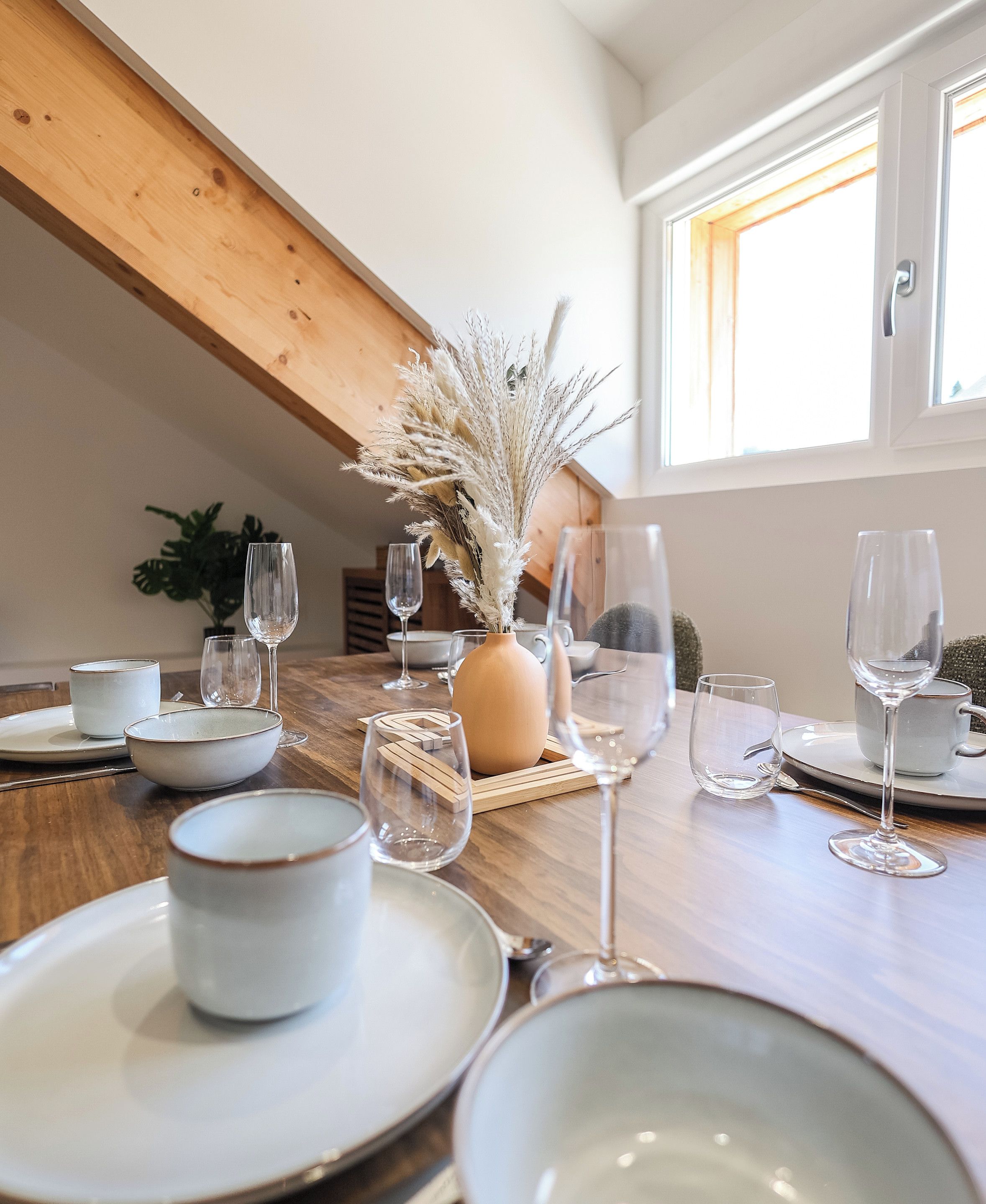 A dining table set with plates, bowls, and wine glasses, with a vase of dried plants in the center.