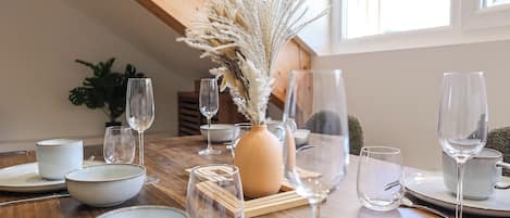 Una mesa de comedor dispuesta con platos, tazones y copas de vino, con un florero de plantas secas en el centro.