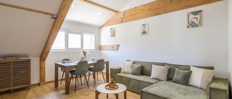 A cozy living room with a wooden ceiling, a green sofa, a dining table with chairs, and a small round table with a vase.