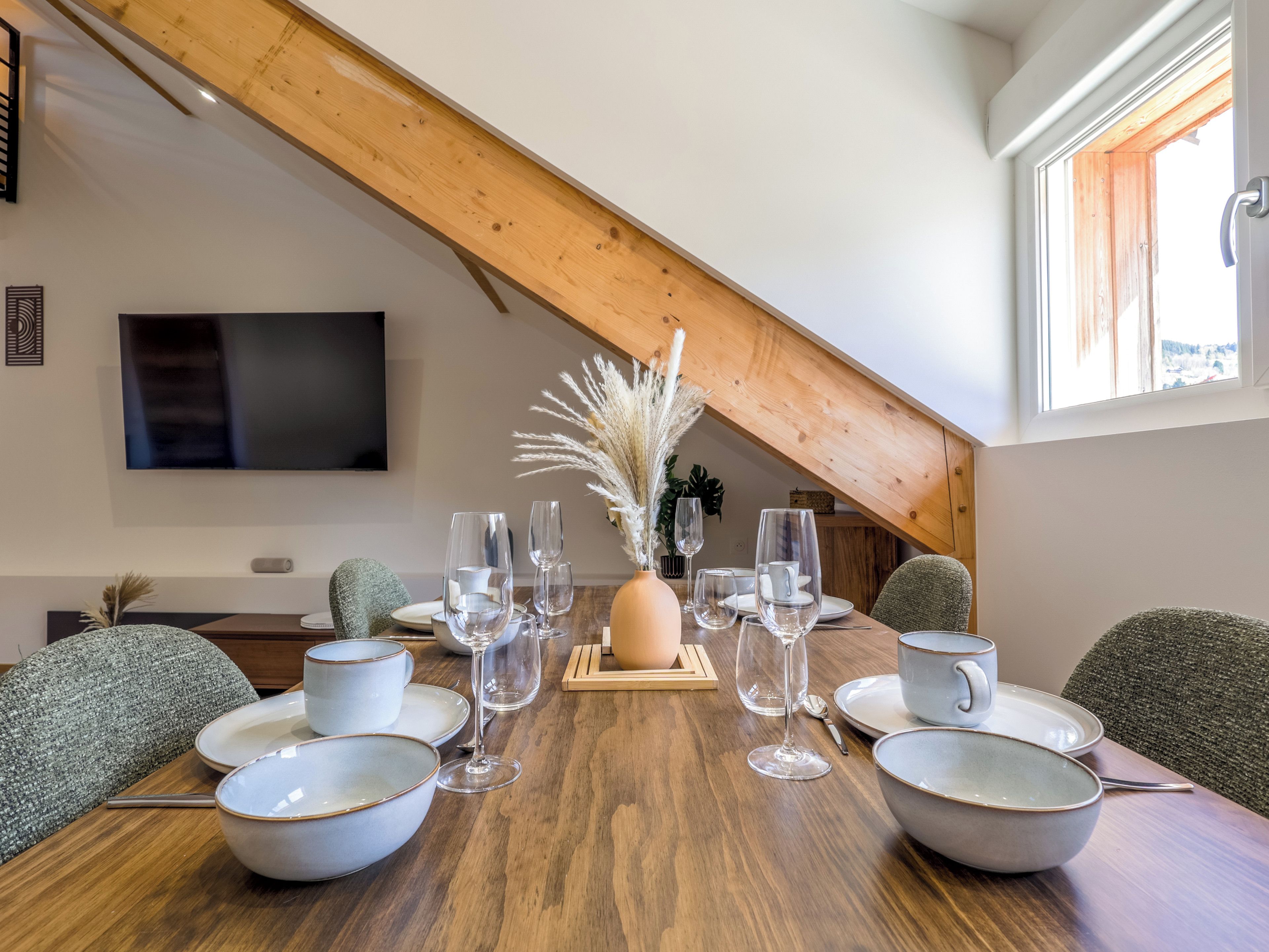 A dining area with a wooden table set for a meal, a flat-screen TV mounted on the wall, and a window with a view.