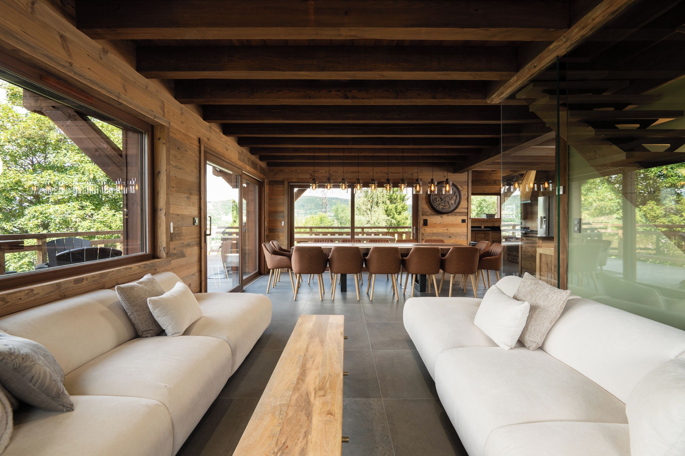 A modern interior with wooden beams, a dining area with chairs, and a large window offering a view of greenery.