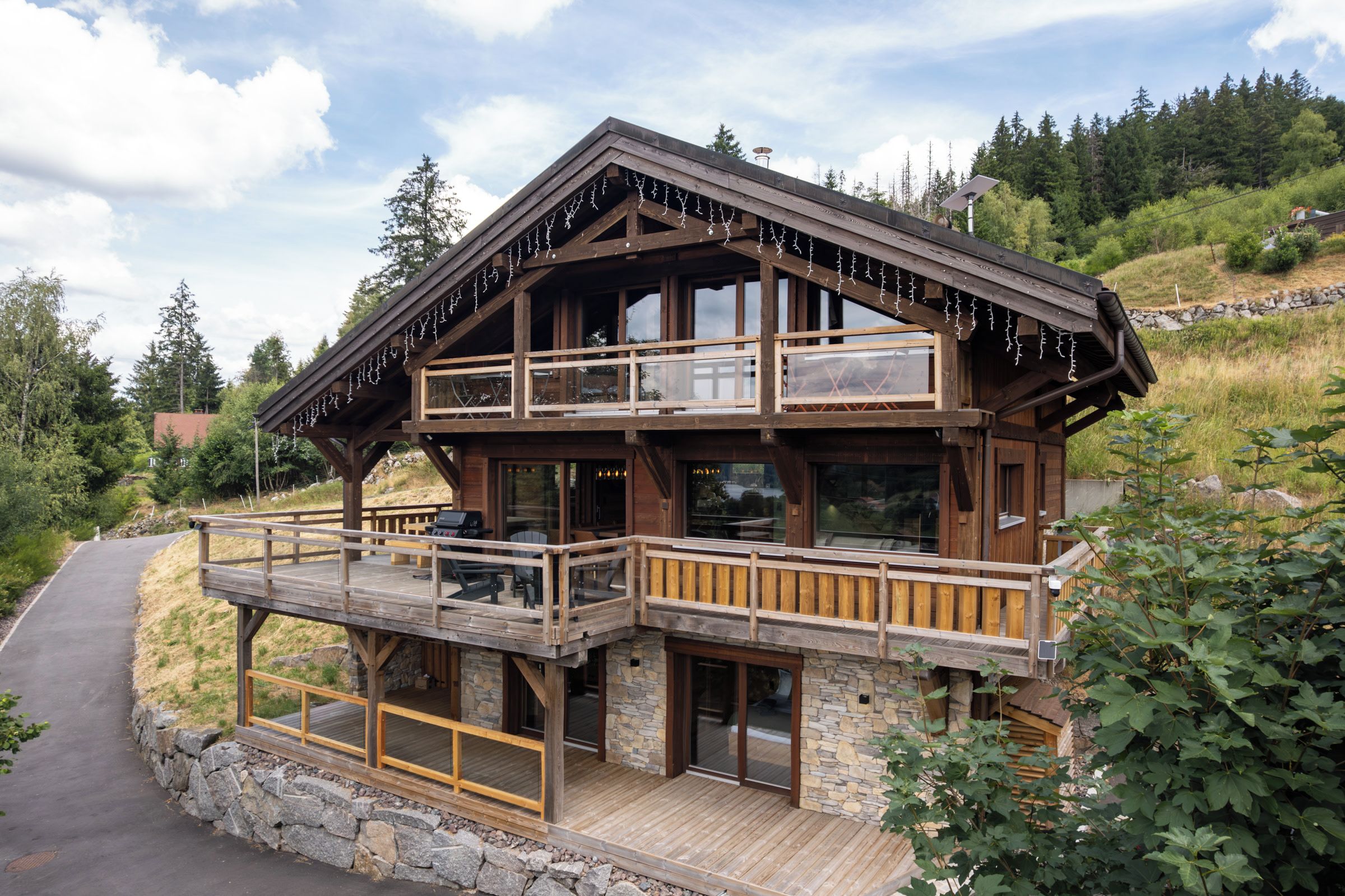 A two-story wooden cabin with a balcony, stone foundation, and a view of a forested hillside.