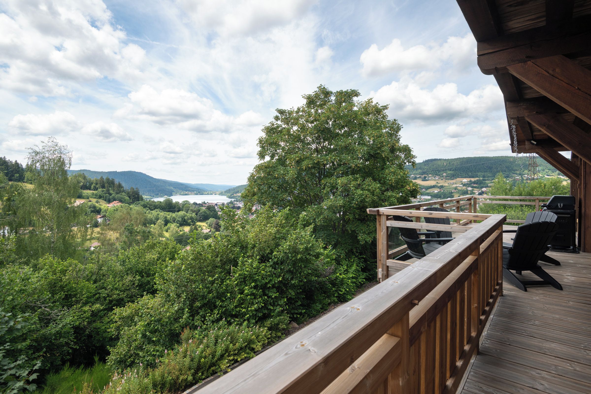 Une terrasse en bois offrant une vue sur un lac, des arbres et des collines lointaines.