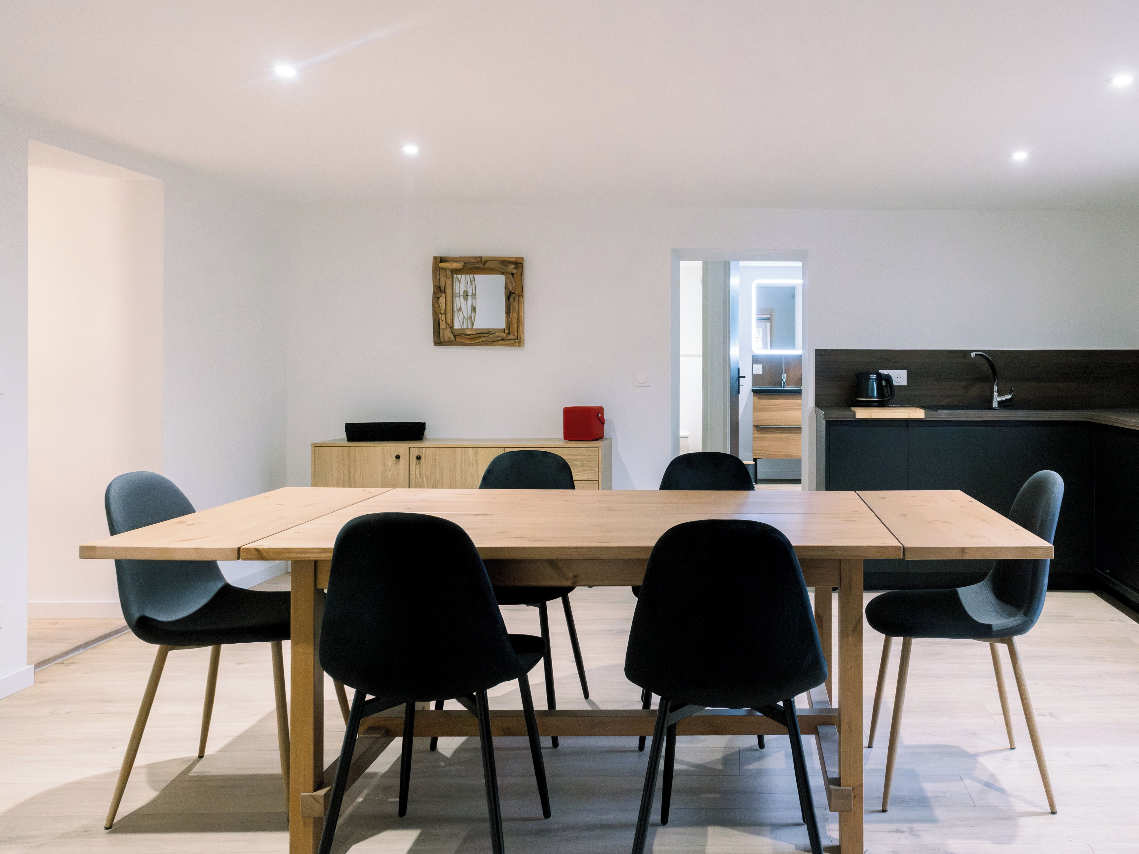 A modern dining area with a wooden table and black chairs, a small cabinet with a mirror, and a kitchen in the background.