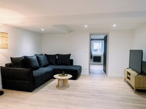 A modern living room with a dark-colored sofa, a small round coffee table, and a television on a wooden stand. - L'Écrin du Haut Pergé ★ Garden • Parking • Netflix • Wi-Fi • 2 Bedrooms (Gérardmer)