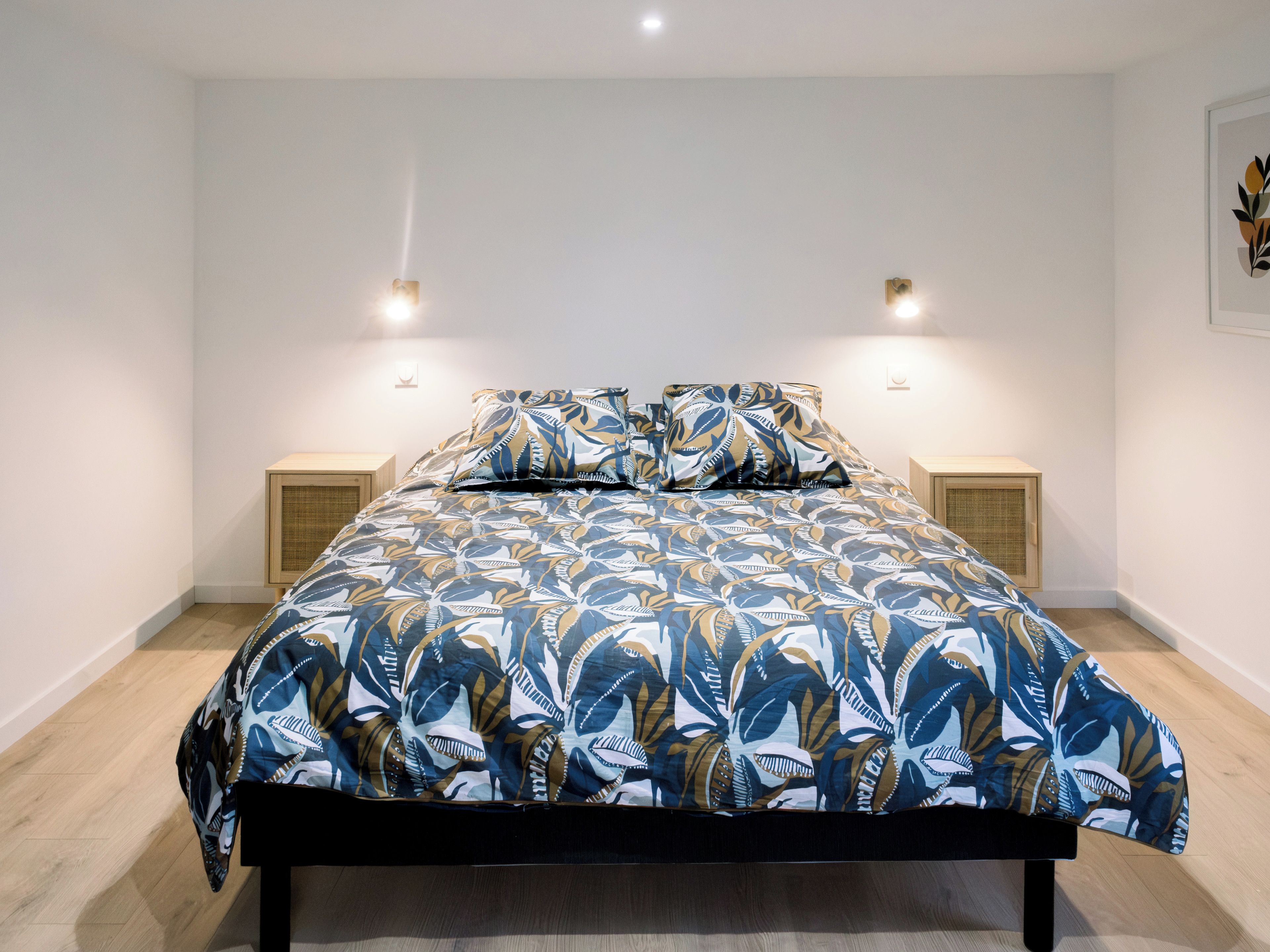 Una cama con una colcha y almohadas estampadas, flanqueada por dos mesitas de noche con lámparas, en una habitación con paredes blancas y suelo de madera.