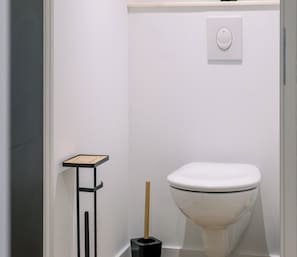 A modern bathroom with a white toilet, a small black trash bin, and a minimalist shelf unit. - L'Écrin du Haut Pergé ★ Garden • Parking • Netflix • Wi-Fi • 2 Bedrooms (Gérardmer)