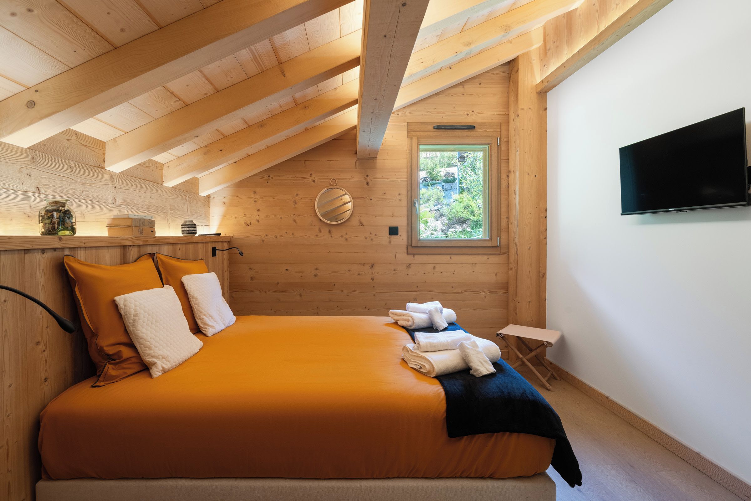 Un dormitorio con techo de madera, cama con ropa de cama naranja, ventana con vistas a la vegetación y un televisor de pantalla plana montado en la pared.