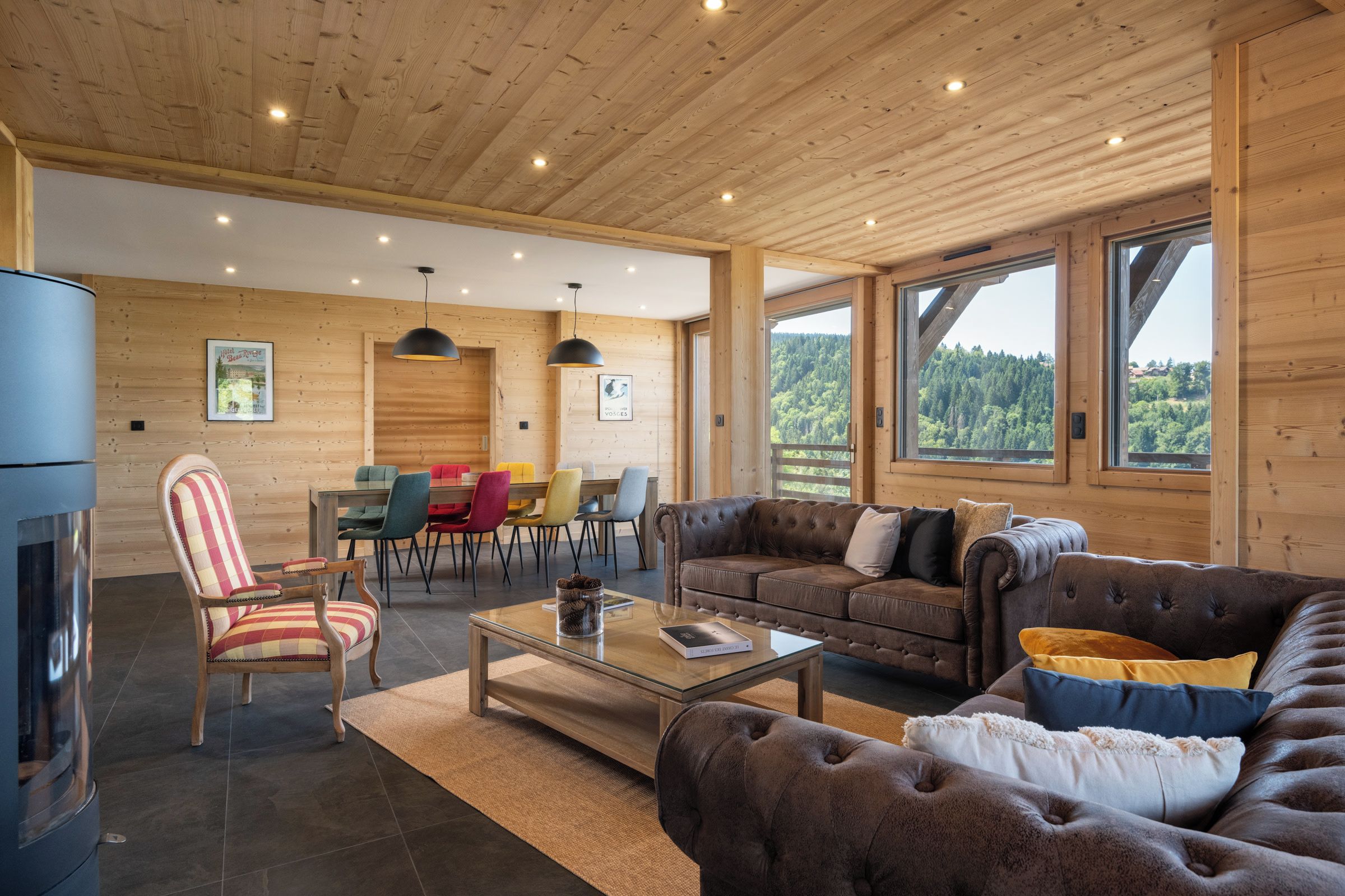 A spacious living area with a fireplace, a dining table with colorful chairs, a sofa with cushions, and large windows offering a view of the forest.