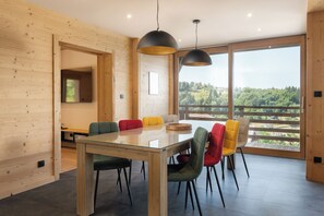 A modern dining area with a glass table, multiple chairs, and large windows offering a view of a forested landscape. - ✨ Chalet Farnes 800 – Brand new Luxury Chalet with Panoramic Lake Views (Gérardmer)