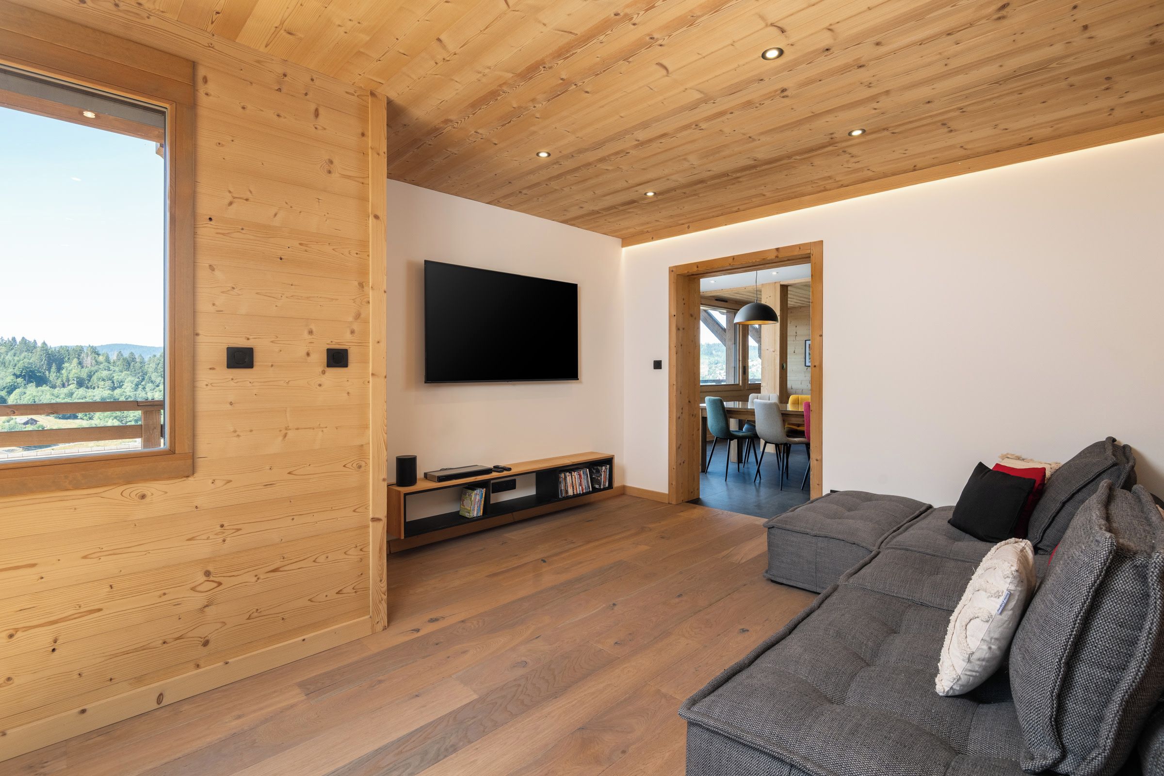 A modern living room with a wooden ceiling, a flat-screen TV mounted on the wall, a sofa with cushions, and a wooden entertainment unit.