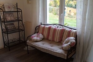 A cozy room with a cushioned bench by a window, a metal shelf with decorative items, and a view of a garden. - Holiday home with 1 bedroom (L'Isle-Jourdain)