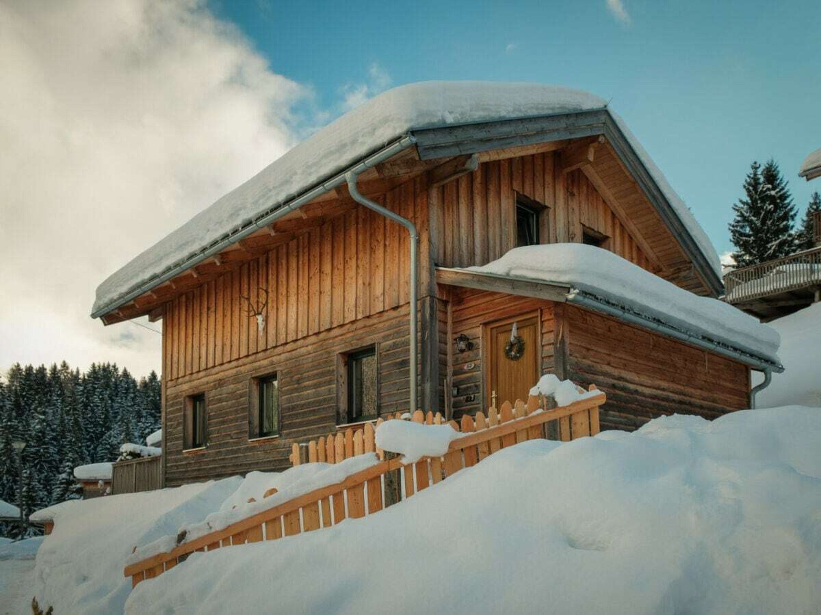 A wooden cabin with a snow-covered roof, a wooden porch, and a wooden fence.
