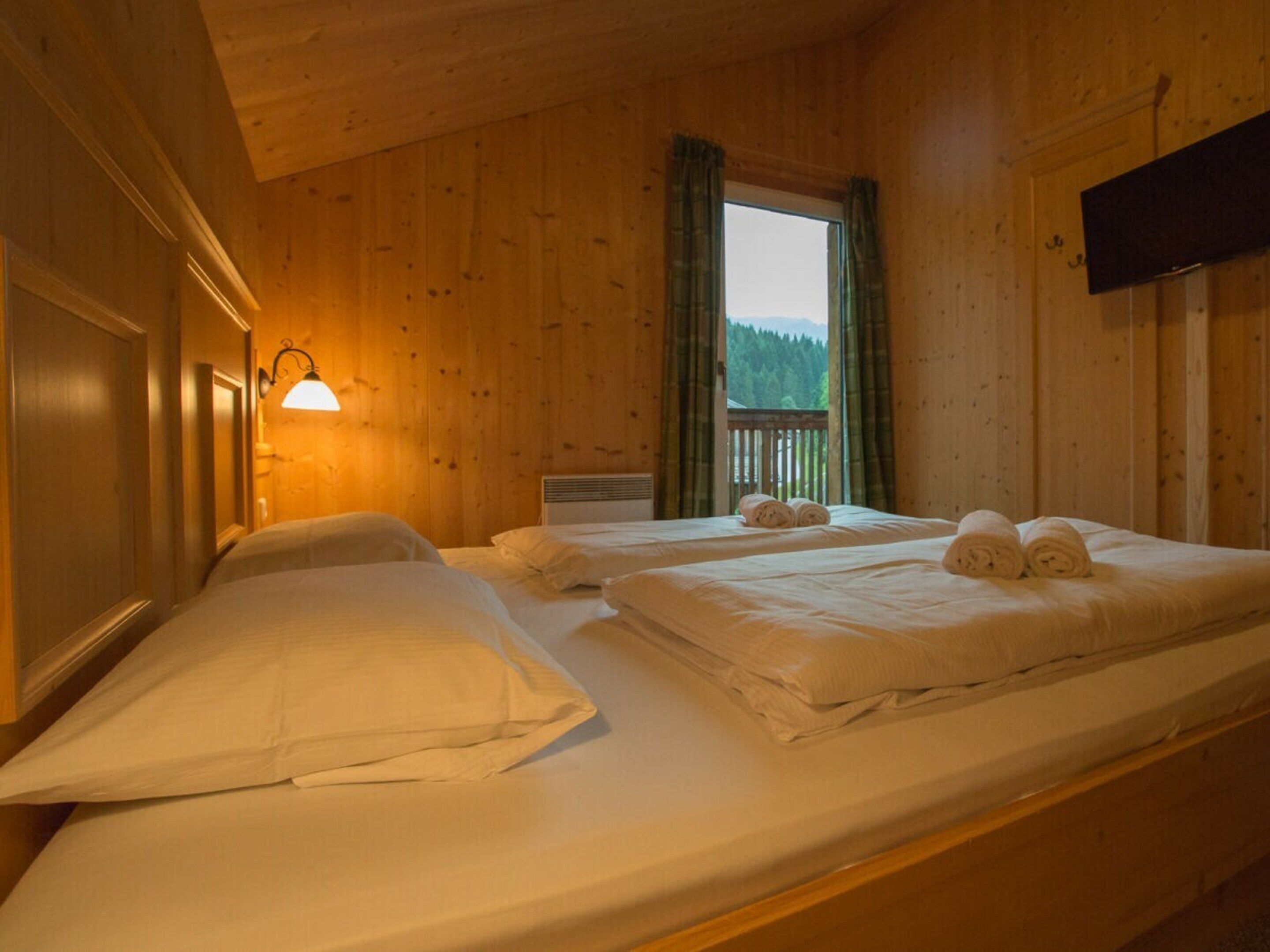 Quarto de cabana de madeira com duas camas, janela com cortinas, televisão montada na parede e abajur fixado na parede.