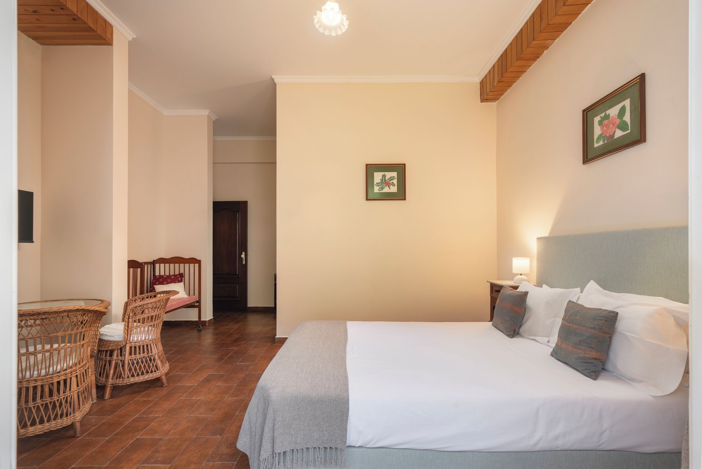 Habitació d'hotel amb un llit, dues taules de nit, cadires de vimetla, una taula petita i una porta que condueix a una altra habitació.