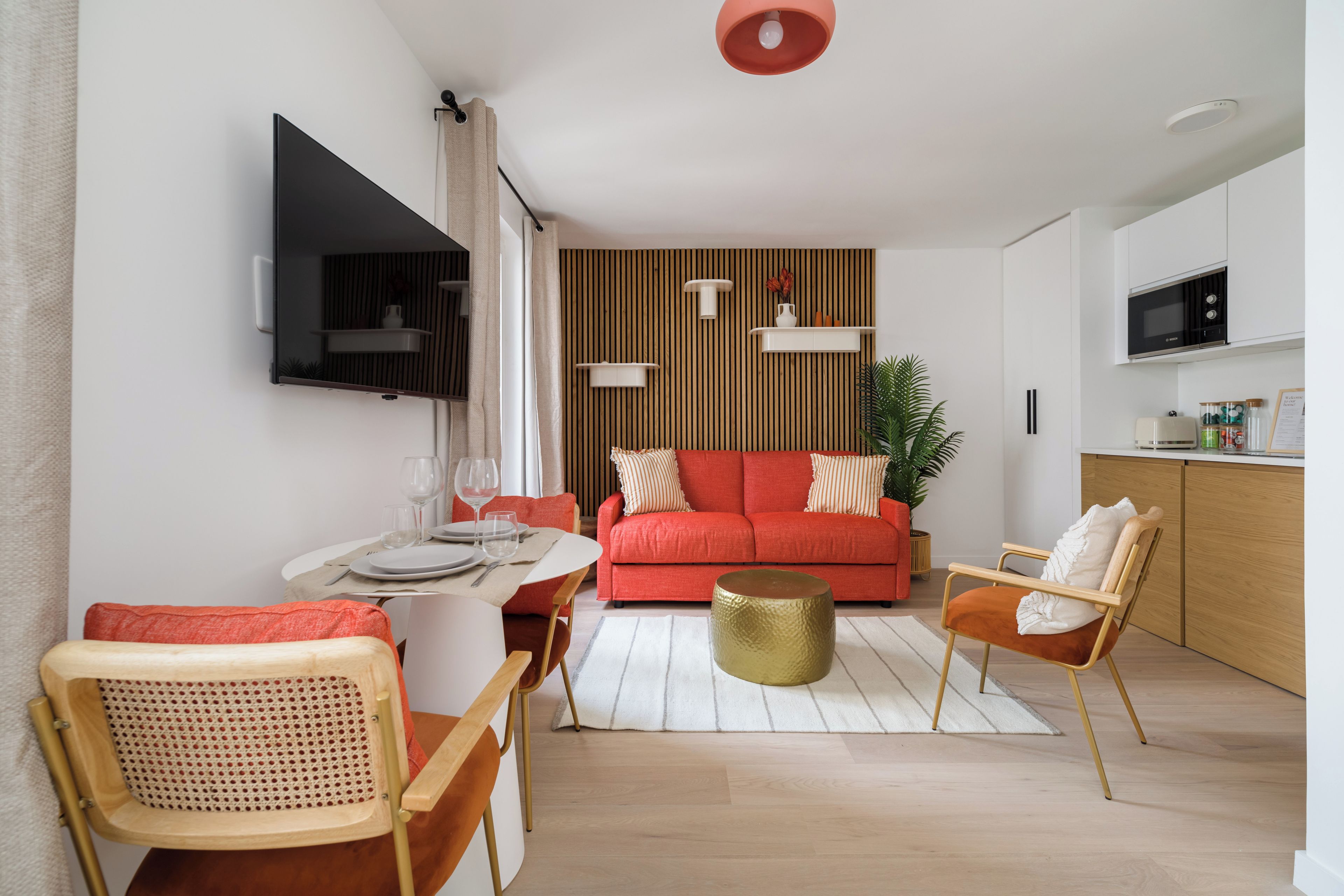 Een moderne woonkamer met een rode bank, houten details, een eethoek met een tafel gedekt voor twee personen, en een keuken met een magnetron.