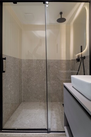 A modern bathroom with a glass shower enclosure, a white sink, and a marble-tiled shower area. - Cozy Studio (Paris)