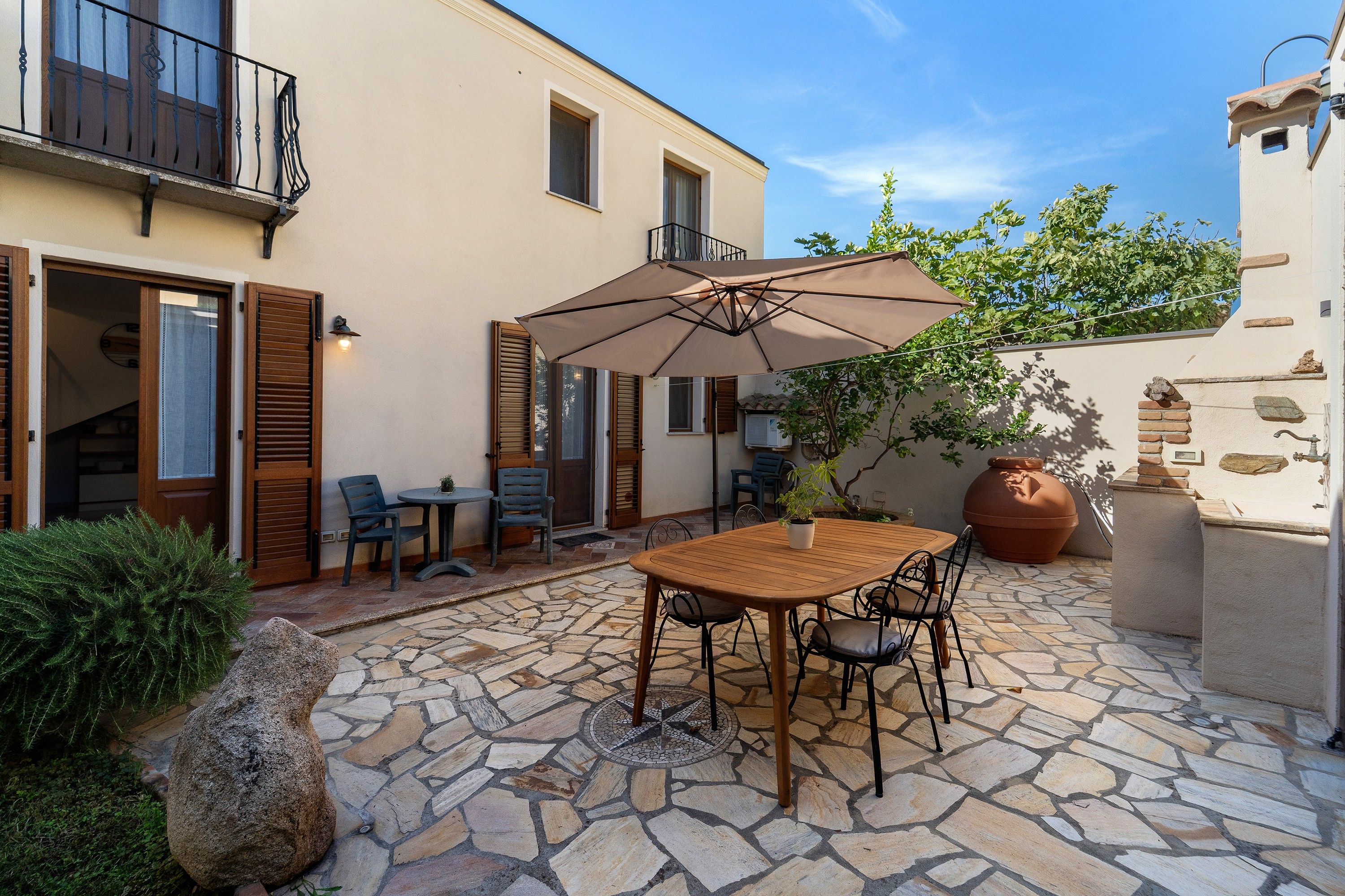 A stone patio with a wooden table and chairs, a large umbrella, and a potted plant.