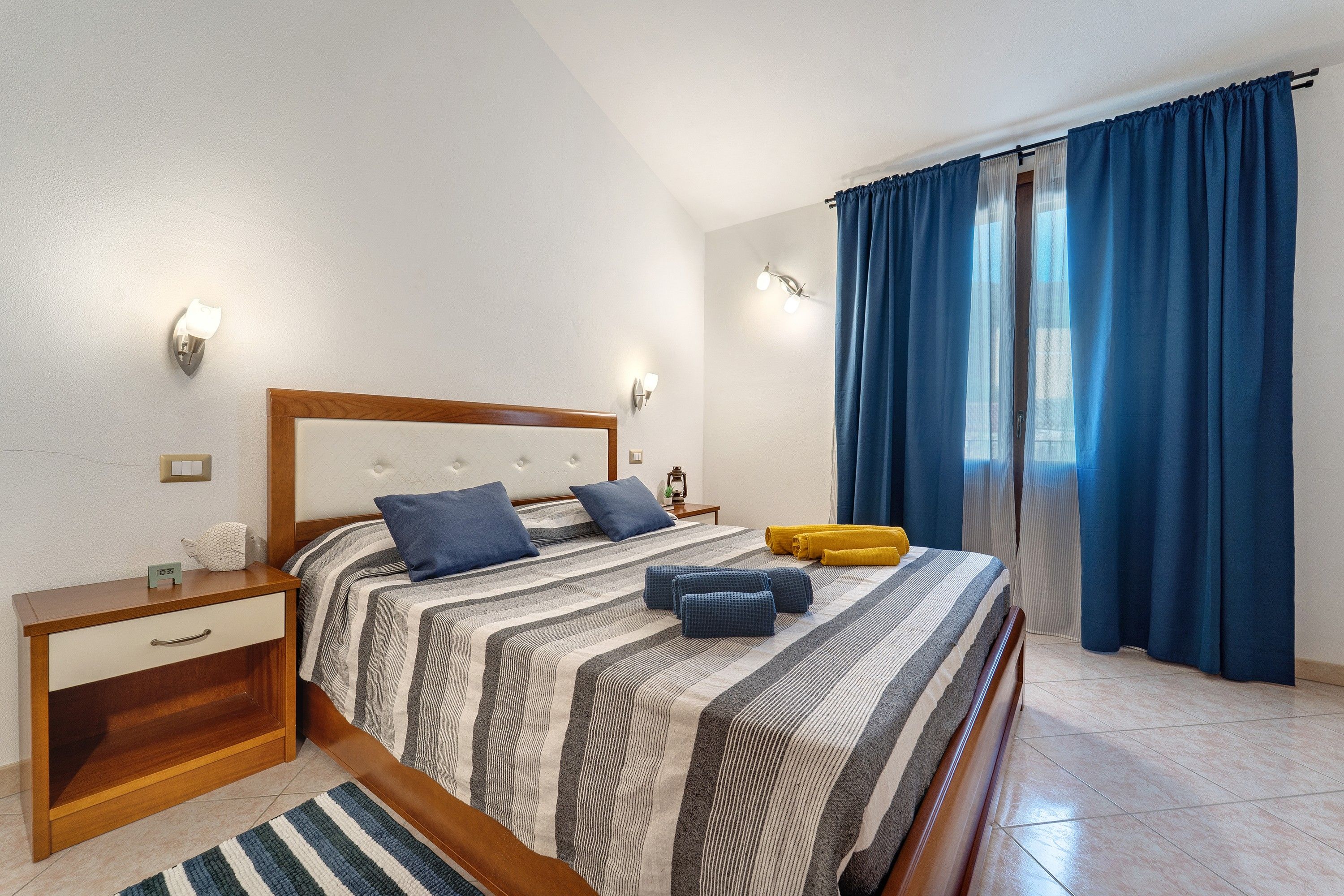 Un dormitorio con cama, mesitas de noche, un reloj de pared y un ventanal con cortinas.