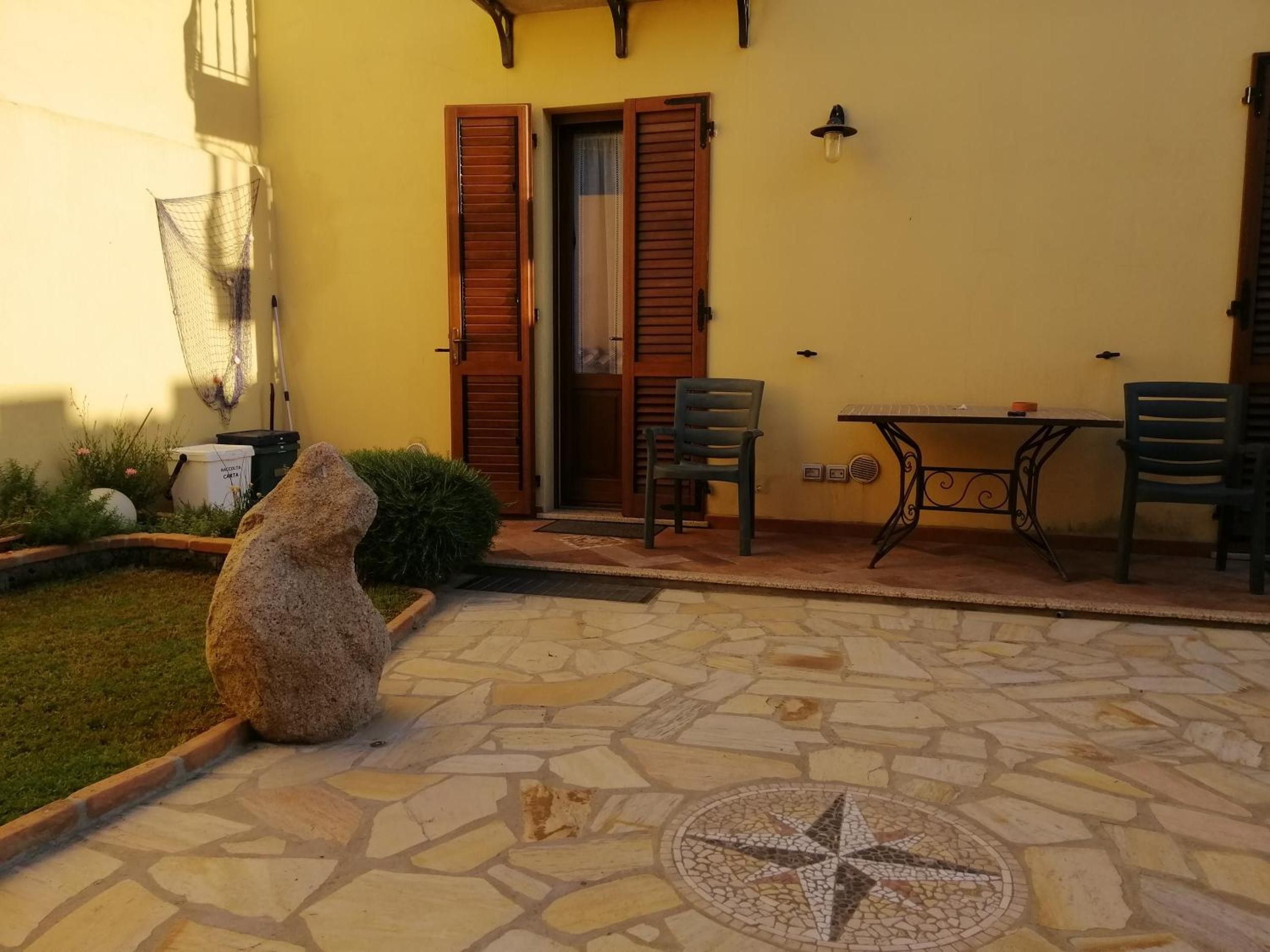 A patio area with a stone floor, a table with chairs, a stone sculpture, and a door with wooden shutters.