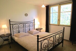 Ein Einzelbett mit Metallgestell und einem blumigen Bettbezug, zwei Nachttische mit Lampen, ein Fenster mit Vorhängen sowie eine Wanduhr.