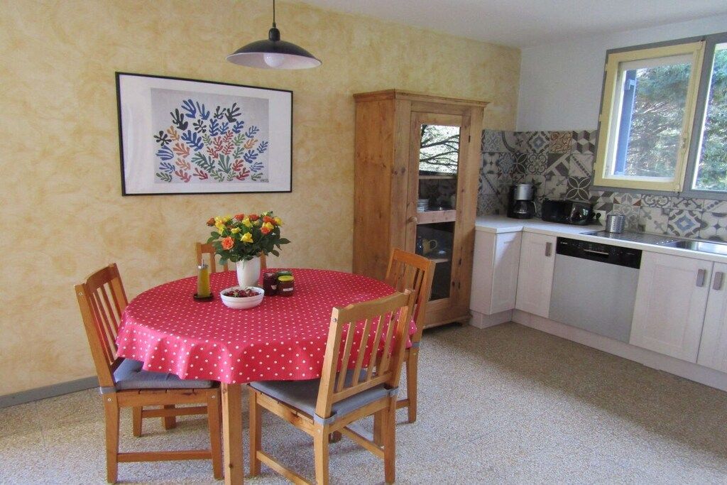 A dining area with a red tablecloth, wooden chairs, a vase with flowers, a framed artwork on the wall, and a kitchen in the background.