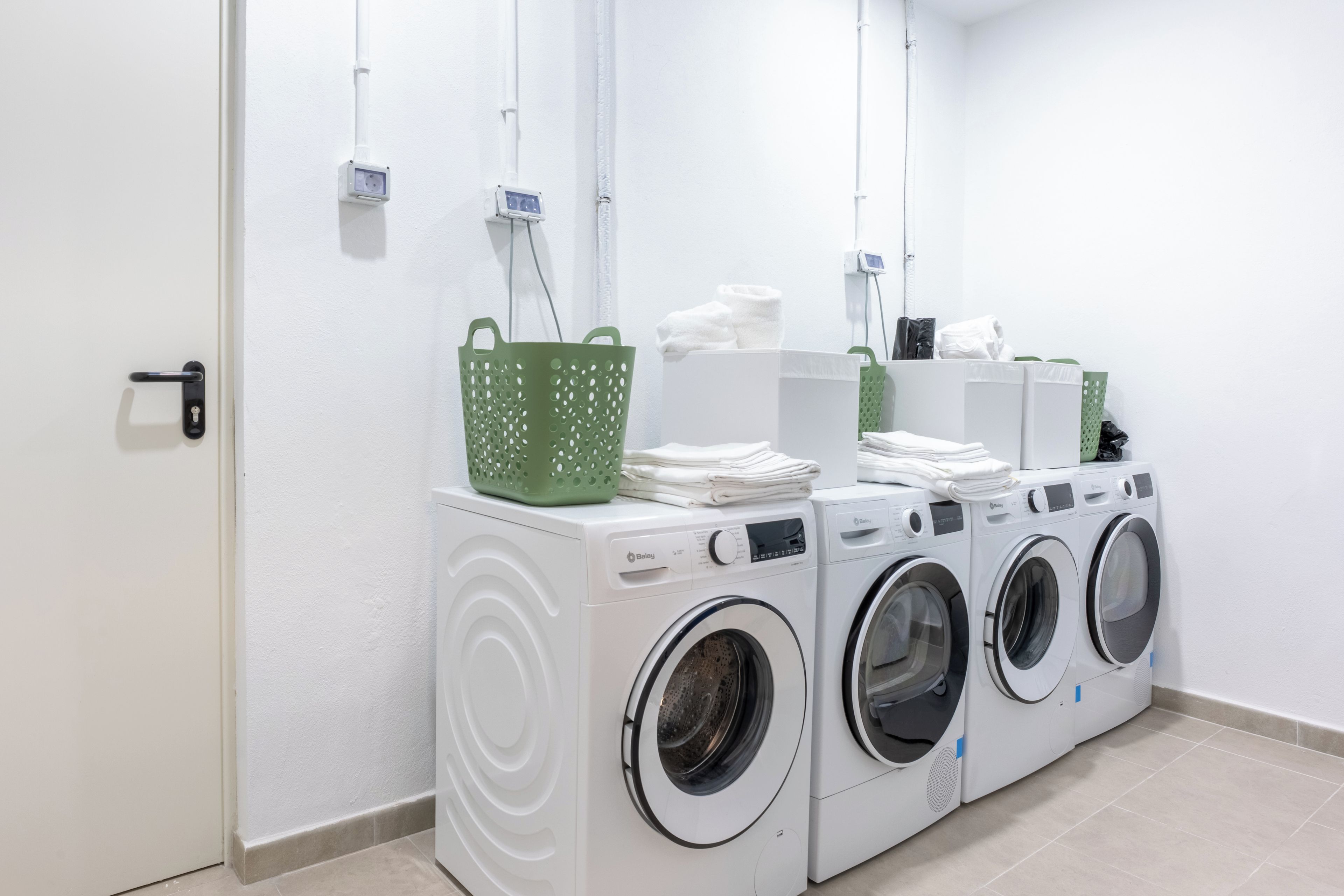 A laundry area with a row of white washing machines, a green basket, and a stack of white towels.
