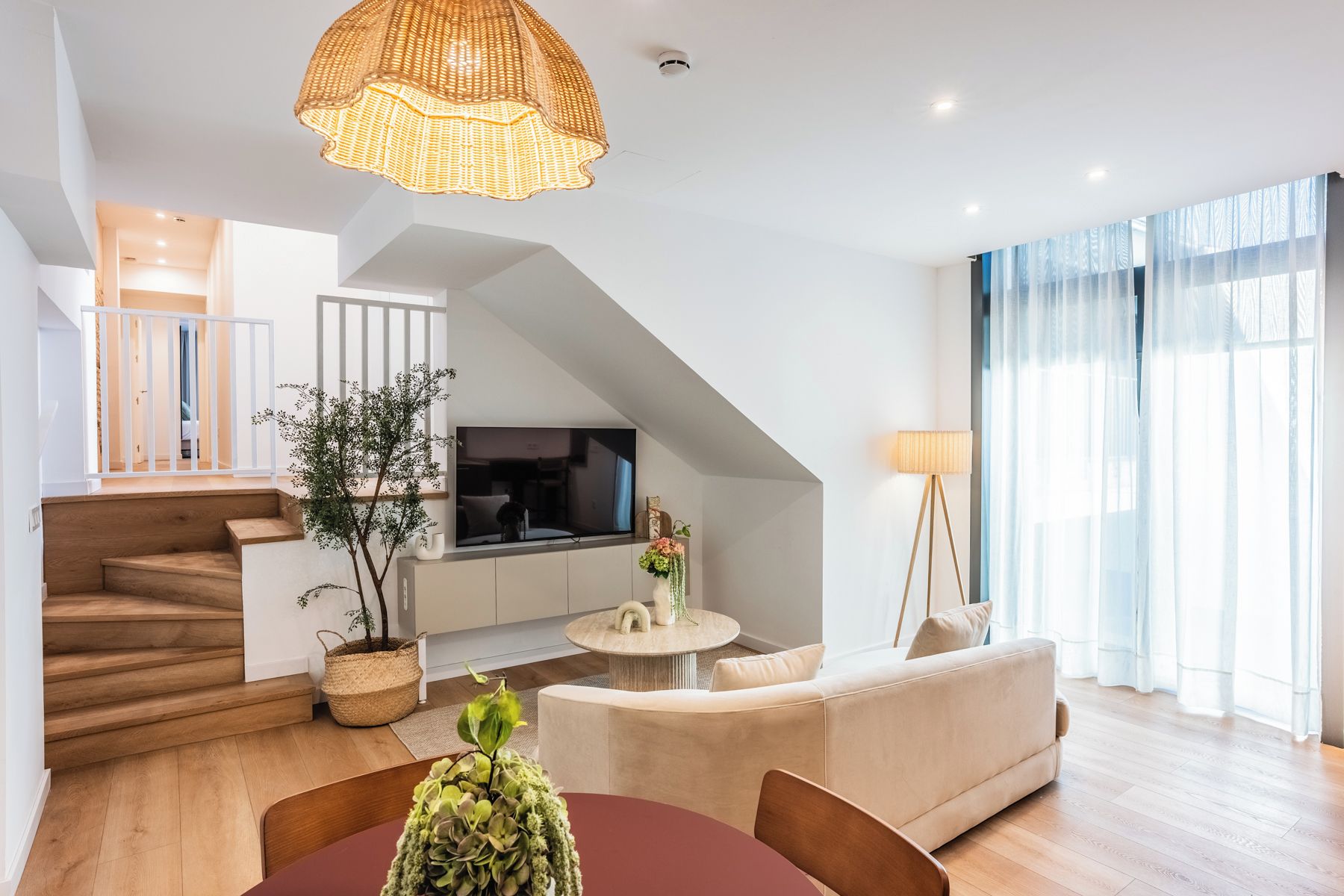 A modern living room with a staircase, a flat-screen TV, a dining area, and a large window with sheer curtains.