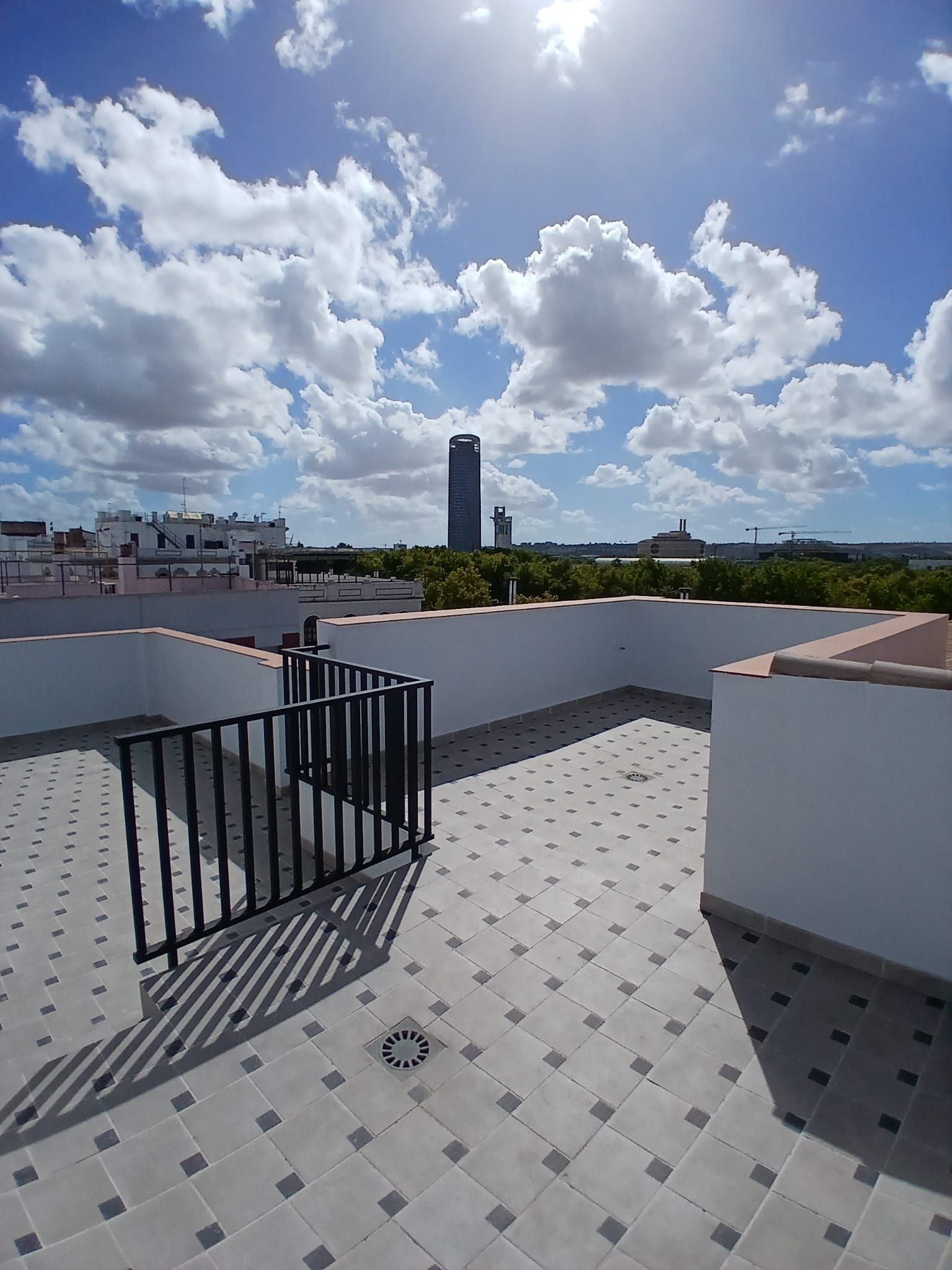 Eine Dachterrasse mit Fliesenboden, Metallgeländer und Blick auf eine Stadtlandschaft, darunter ein hohes Gebäude.