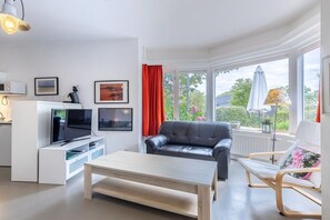 Ein modernes Wohnzimmer mit Flachbildfernseher, Ledersofa, Couchtisch aus Holz und Fenster mit Blick auf Grünflächen.