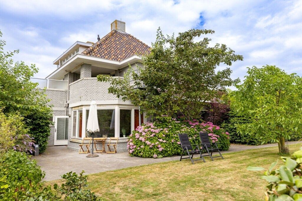 A two-story house with a covered patio area, surrounded by lush greenery and flowering plants.