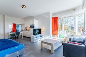 Ein modernes Wohnzimmer mit einer Couch, einem Couchtisch und einem Fernseher. Es gibt eine Küchenzeile mit einer Mikrowelle und einem Esstisch. Das Zimmer verfügt über große Fenster mit Vorhängen.