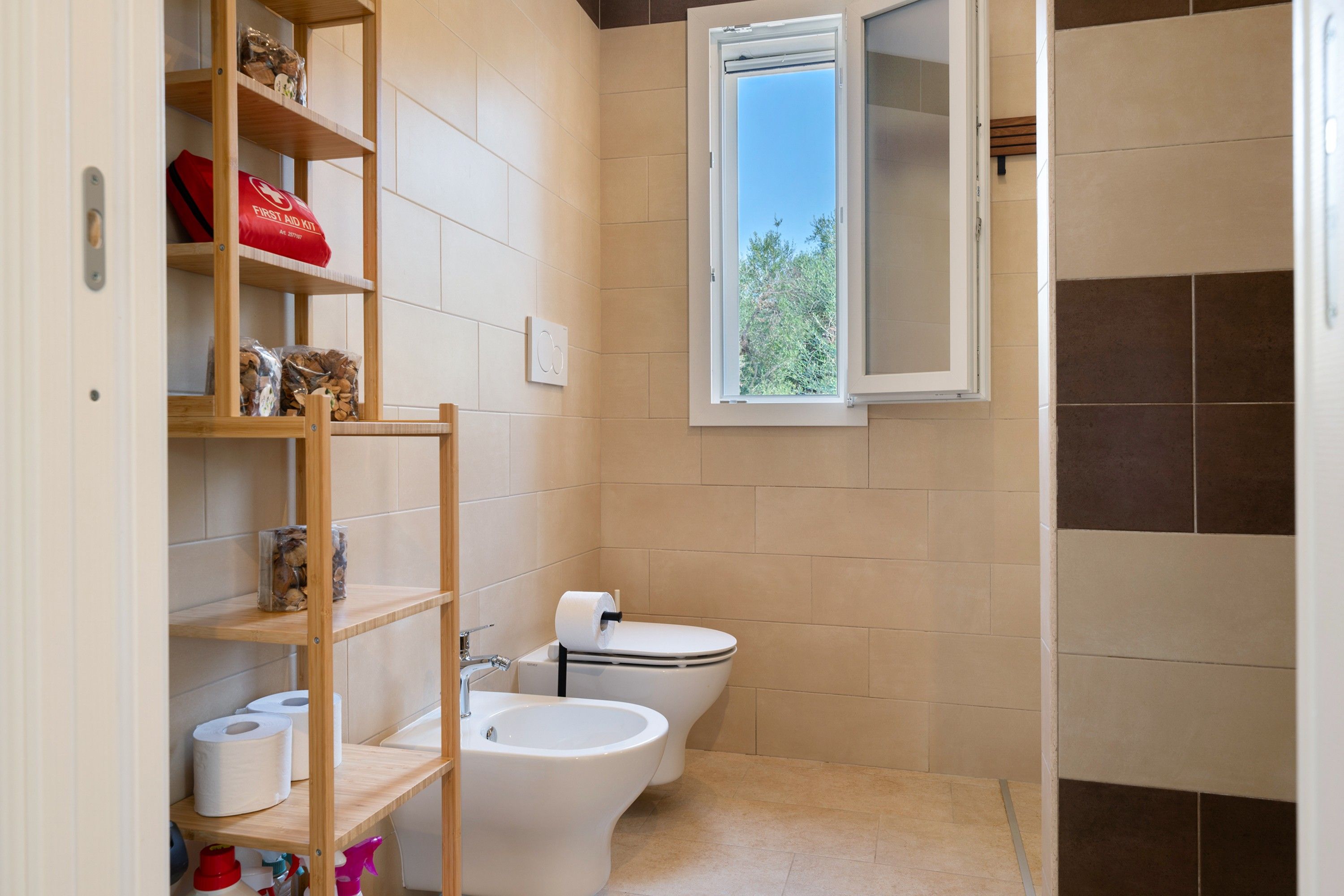Une petite salle de bain comprenant des toilettes, un bidet, des étagères avec des articles de toilette et une fenêtre.