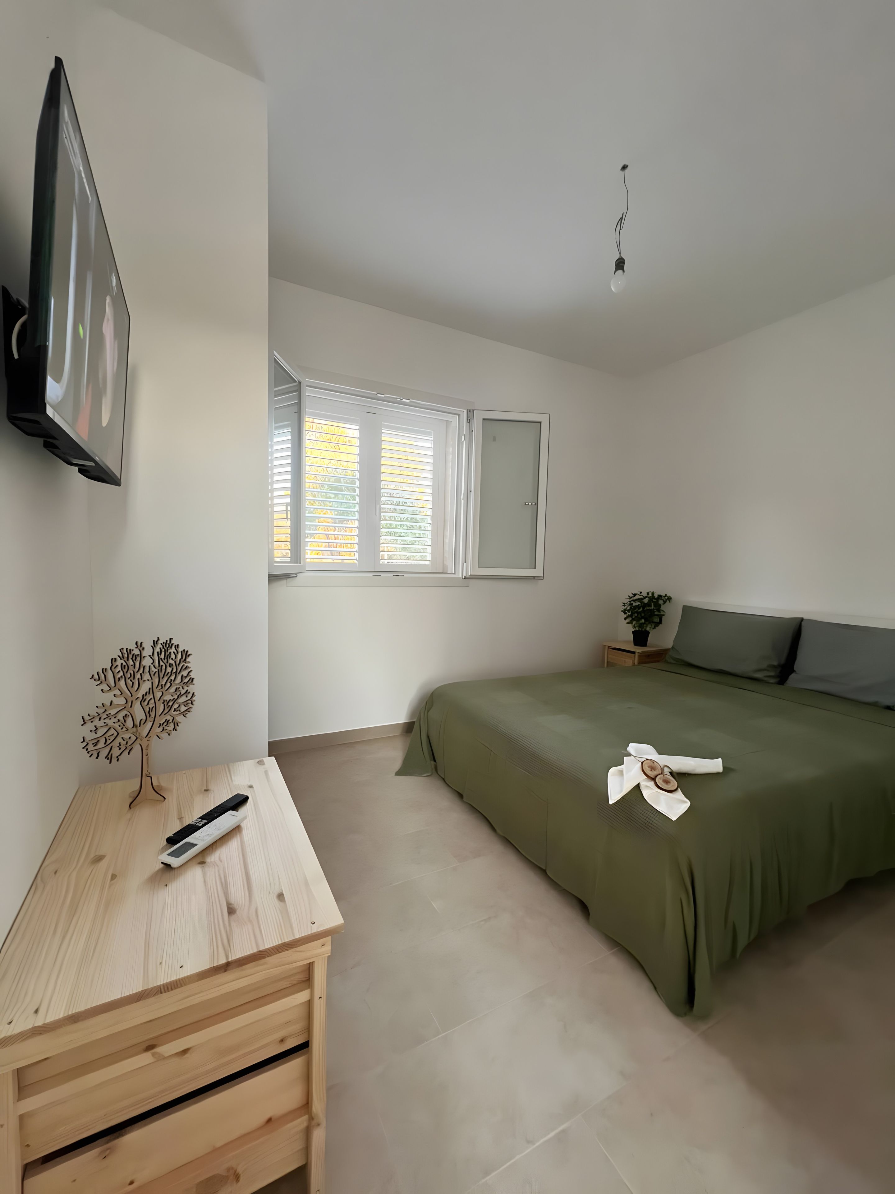 Camera da letto minimalista con un letto, una TV a parete, un comodino in legno e una finestra con persiane.