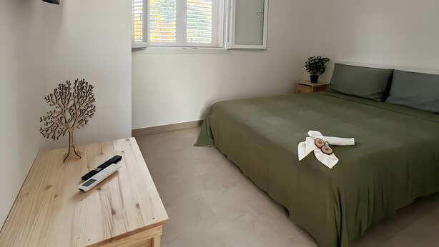 Quarto minimalista com uma cama, televisão presa à parede, mesa de cabeceira de madeira e janela com persianas.