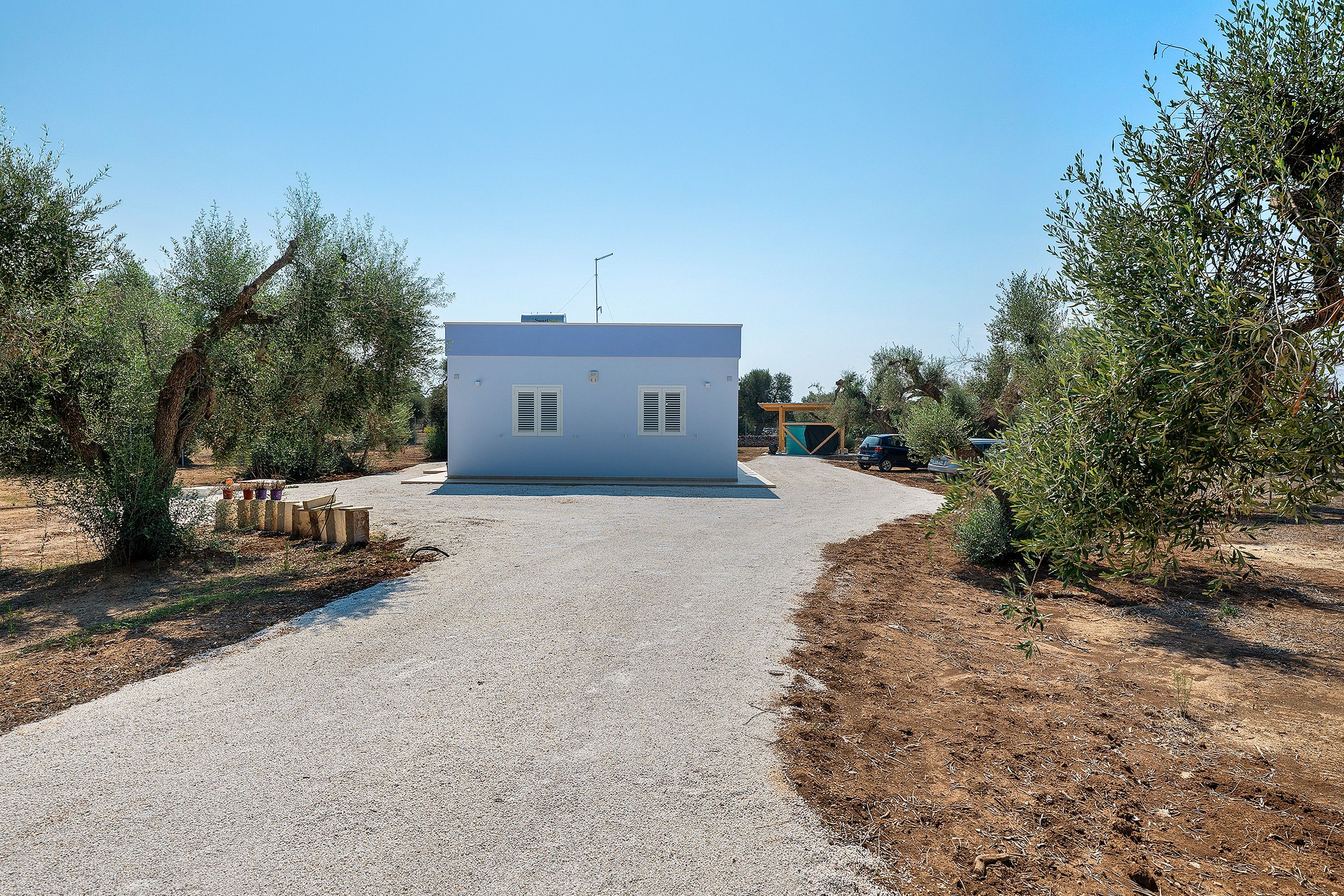 Un petit bâtiment d’un seul étage, avec une allée de gravier menant à l’entrée, entouré d’oliviers.