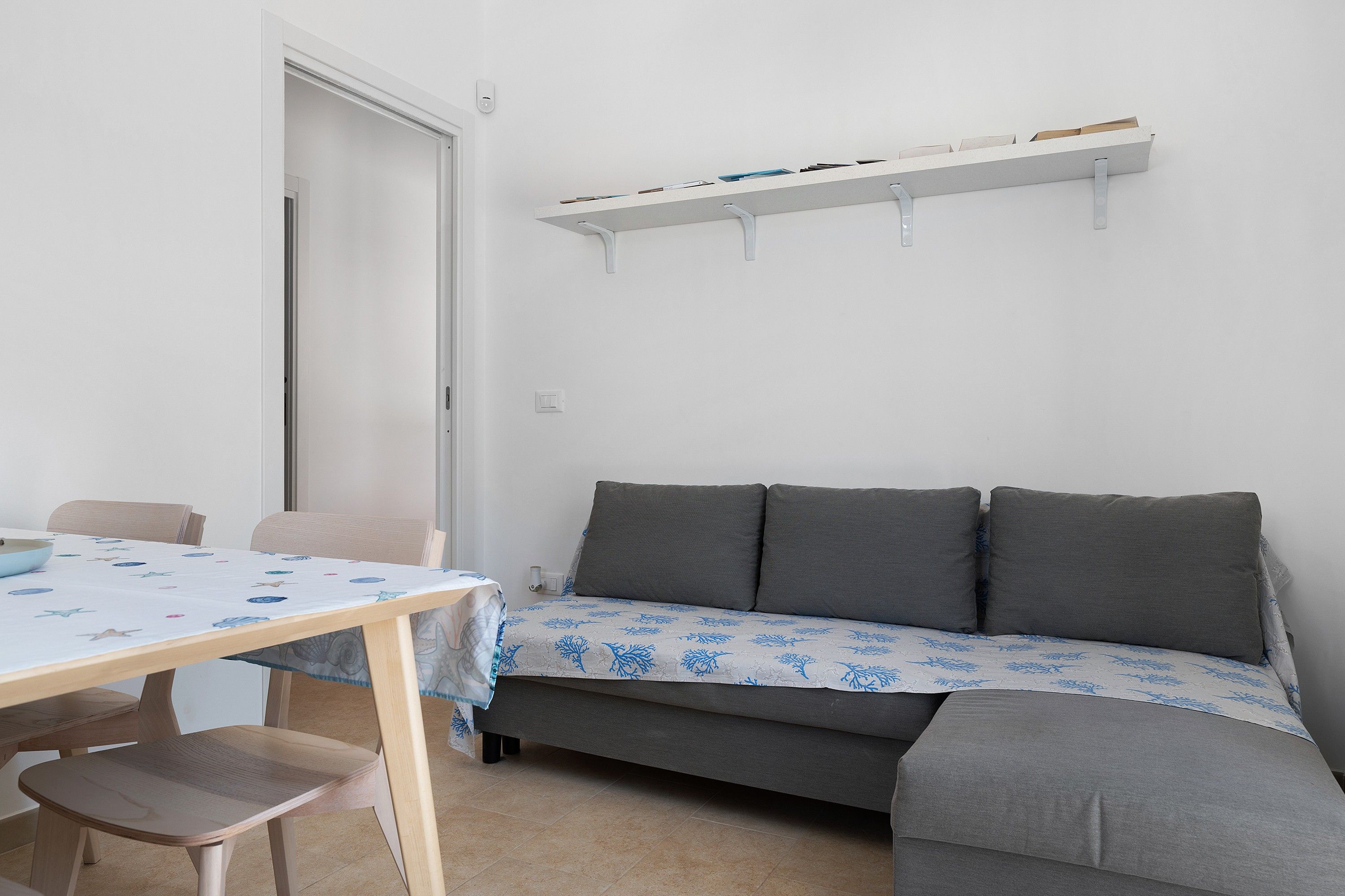 Ein Zimmer mit einer Couch, einem Tisch und Stühlen. Eine Tür und ein Bücherregal sind ebenfalls vorhanden.