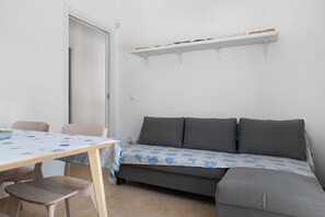 A room with a sofa, a table, and chairs. There is a door and a shelf with books.
