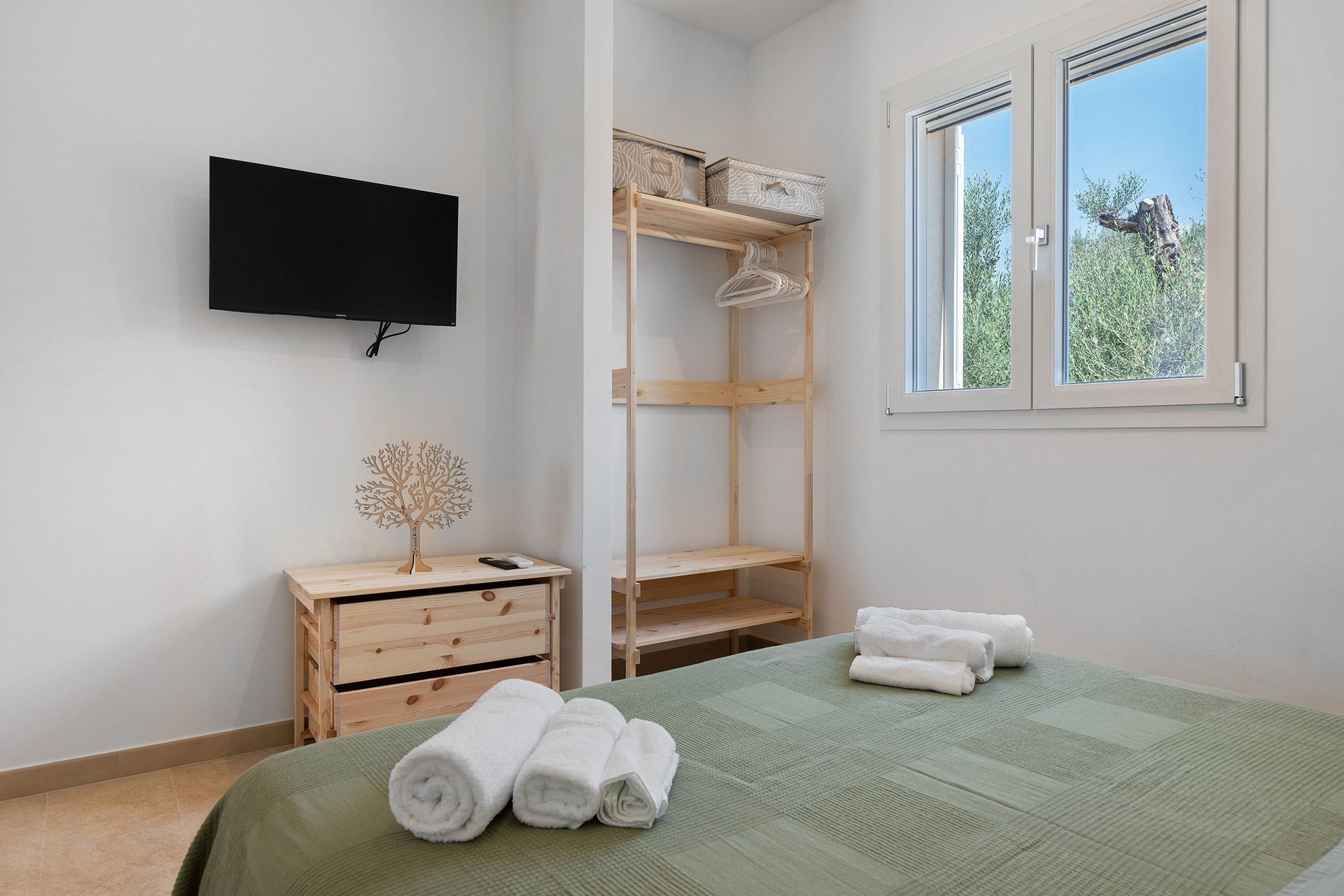 Ein Schlafzimmer mit einem Bett, einem an der Wand befestigten Fernseher, einer Holzkommode, einem Regalsystem und einem Fenster mit Blick auf Grünflächen.