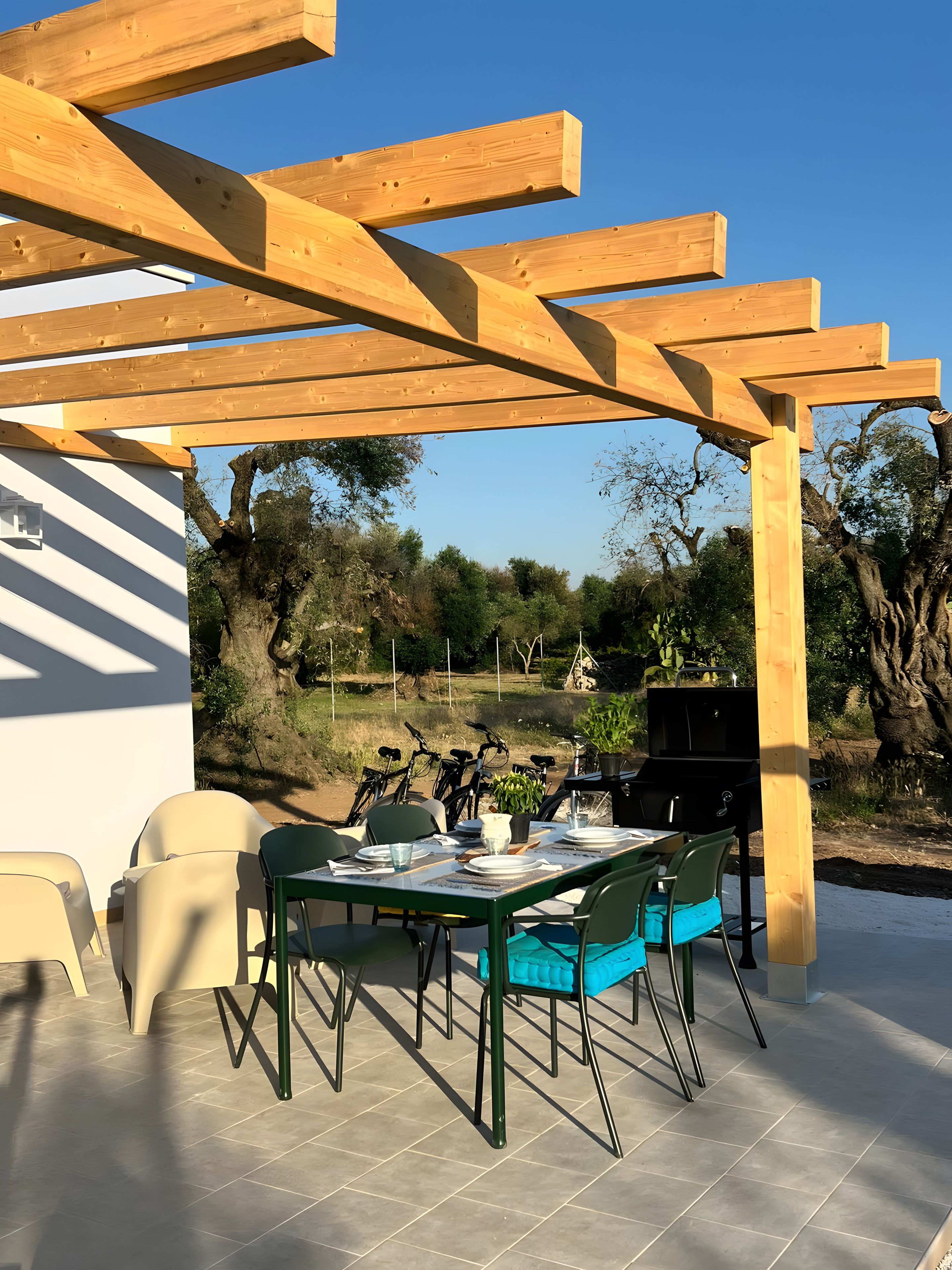 Patio esterno con una pergola in legno, un tavolo da pranzo con sedie, un barbecue e biciclette sullo sfondo.