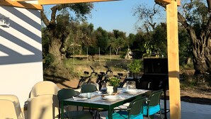 Um pátio externo com pérgola de madeira, mesa de jantar com cadeiras, churrasqueira e bicicletas ao fundo.