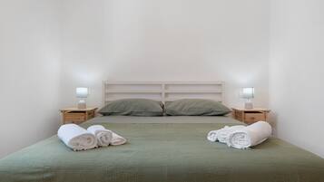 Ein ordentlich bezogenes Bett mit grüner Bettwäsche und zwei Kissen, zwei Nachttische mit Lampen und zusammengerollte Handtücher auf dem Bett.