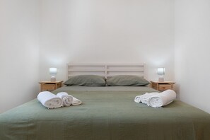 A neatly made bed with green bedding and two pillows, two bedside tables with lamps, and rolled towels on the bed.