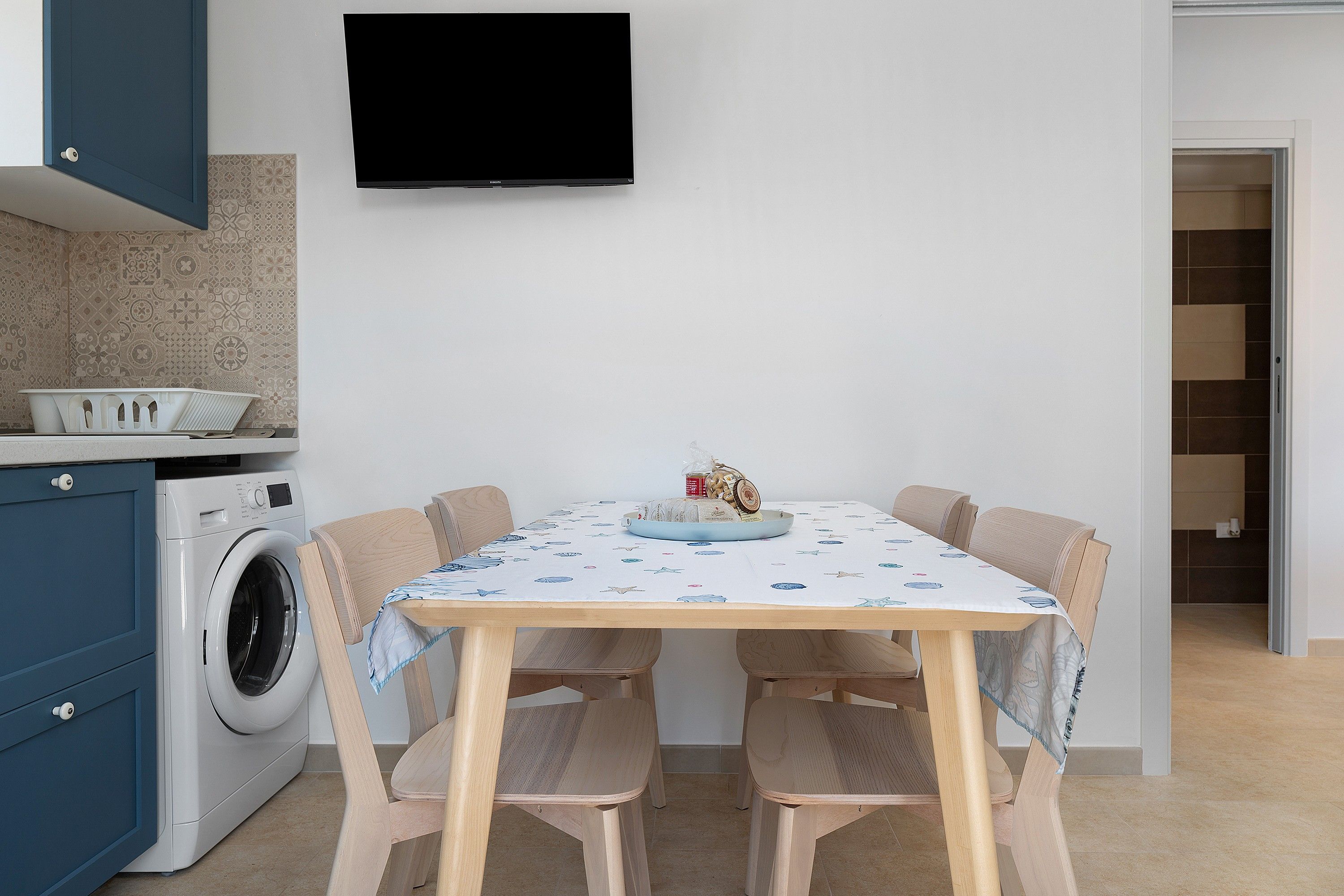Une cuisine moderne équipée d’un lave-linge, d’une table à manger avec des chaises et d’un téléviseur à écran plat fixé au mur.