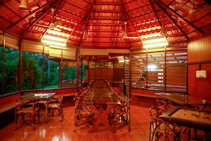 Un vaste espace intérieur doté d’une longue table à manger entourée de chaises, de grandes fenêtres et d’un haut plafond recouvert d’un toit en métal ondulé.
