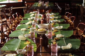 A long dining table set with traditional Indian meal arrangements, including banana leaves, various dishes, and glasses.