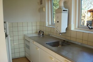 A kitchen with white cabinets, a stainless steel countertop, a built-in oven, a sink, and a window with a view of trees. - Holiday home with 1 bedroom (Callantsoog)