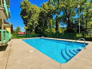 Ein rechteckiger Pool mit Metallleiter und Trepfen, umgeben von einem grünen Zaun, mit Bäumen und einem Gebäude im Hintergrund.