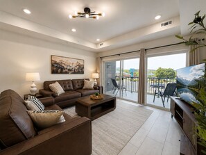 A modern living room with a brown leather sofa, a coffee table, and a flat-screen TV. There is a large window with a view of a parking lot and greenery outside. - New Pige Forge Condo | Mtn View - Walk Everywhere (Pigeon Forge)